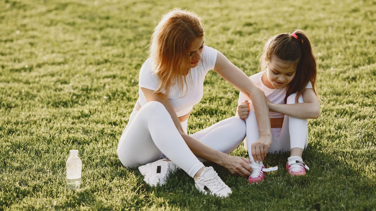 A mother and daughter in activewear bond outdoors on a sunny day, tying shoelaces on the grass.